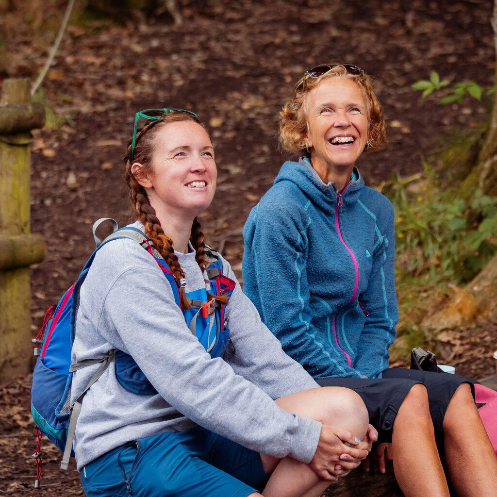 Two people in outdoor clothing, sat in woodland and smiling at something in the trees