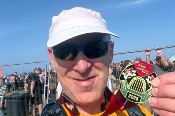 A selfie of Bendrigg's Great North Run fundraisers, Eamonn. Eamonn is a white man with short facial stubble, wearing a white baseball cap, black sunglasses, and a yellow Bendrigg t-shirt. He is smiling into the camera and holding up his Great North Run 2026 medal. Groups of people gather on the grassy area in the background.