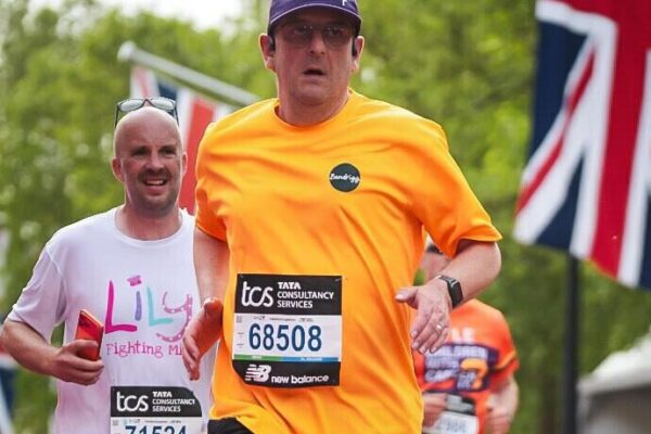 A runner mid-stride at the London Marathon with other runners in the background. He is wearing a yellow t-shirt with a green Bendrigg logo on it, black shorts, and a navy blue baseball cap. He has a calm but determined look on his face.