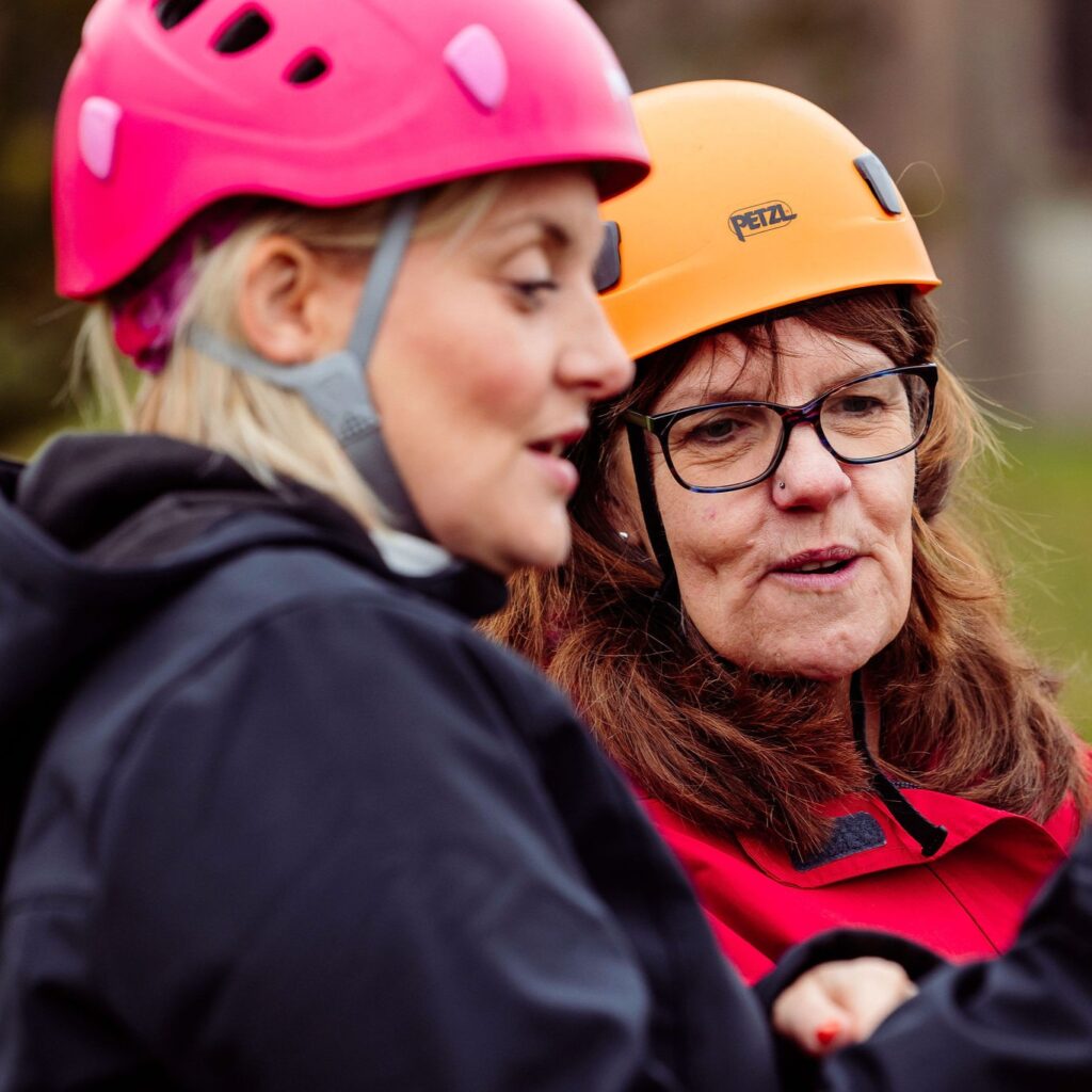 Two people wearing climbing helmets in the outdoors, talking to each other