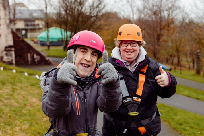 2 people with the thumbs up, smiling. wearing helmets and harnesses