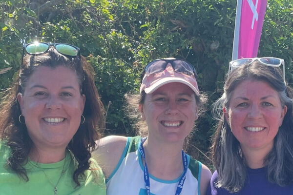 Three women standing together smiling. They are outside on a sunny day.