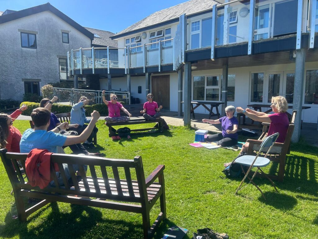 A group of people sit in a circle outside on benches outside on a grassy lawn. They are all making signs with their hands.
