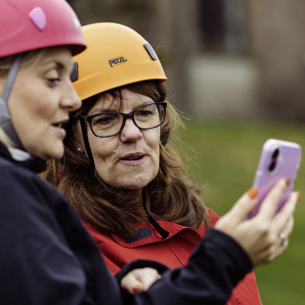Close-up of two people wearing climbing helmets and outdoor clothing, viewing a smartphone screen while standing outside.