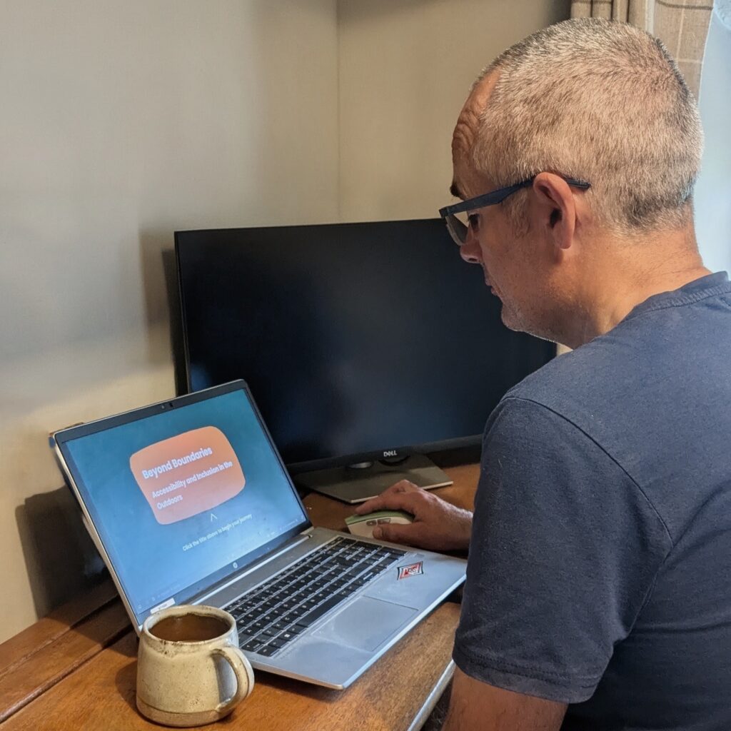 Person sitting at a desk using a laptop and mouse. The laptop screen displays a presentation titled "Beyond Boundaries: Accessibility and Inclusion in the Outdoors".