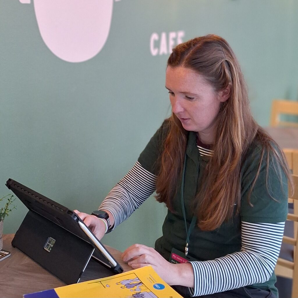 Person sitting at a table and interacting with a tablet in a café, with books and a hot drink visible on the table.